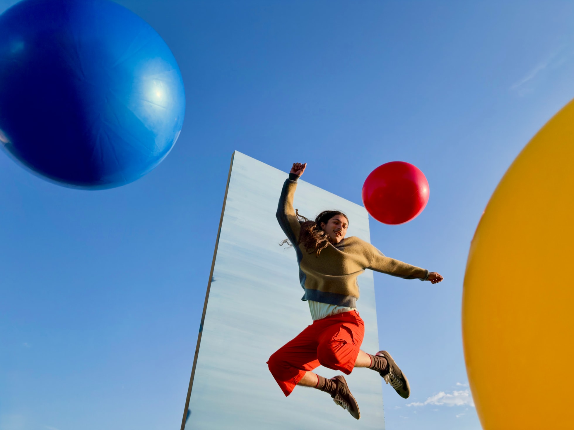 Outdoors, a person jumps as colorful inflated balls are tossed in the air against a blue sky and a rectangular gray backdrop. — Picture from apple.com