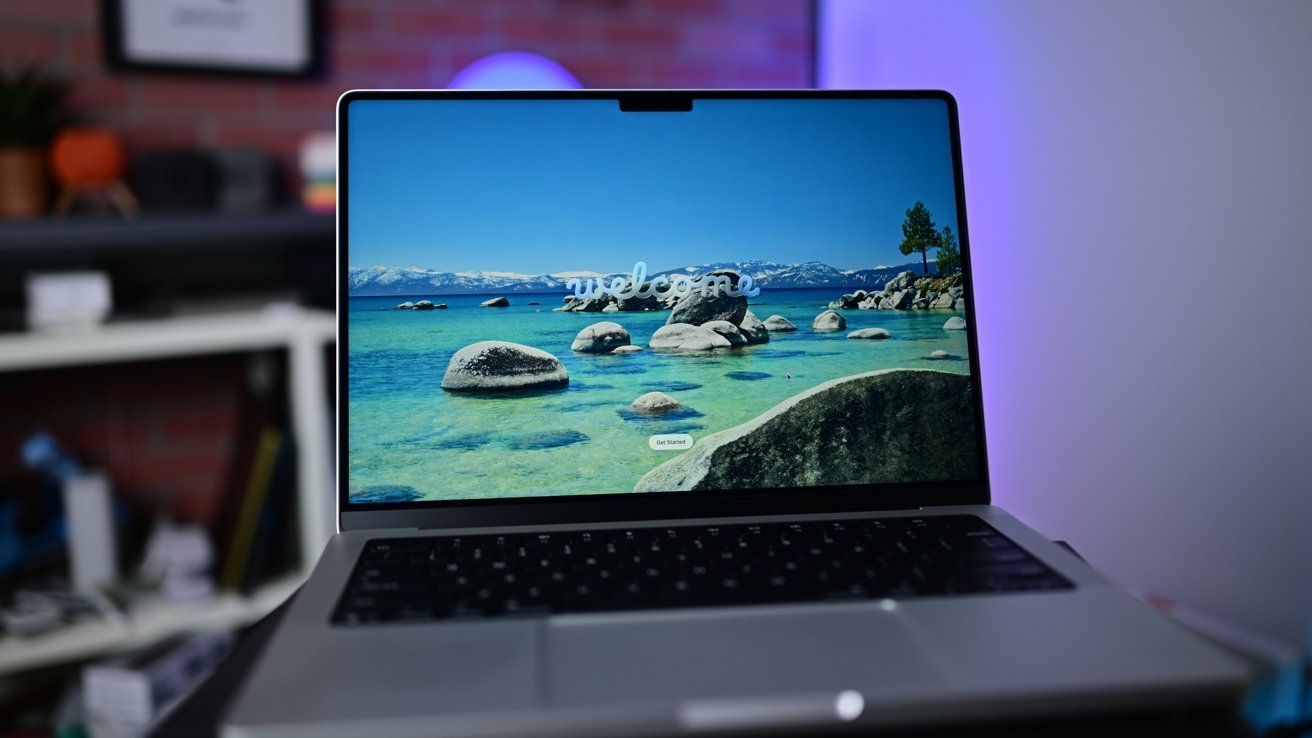Open laptop displaying a vivid beach scene with rocks in clear blue water, set on a desk in a softly lit room with blurred shelves and brick wall in the background — Picture from f