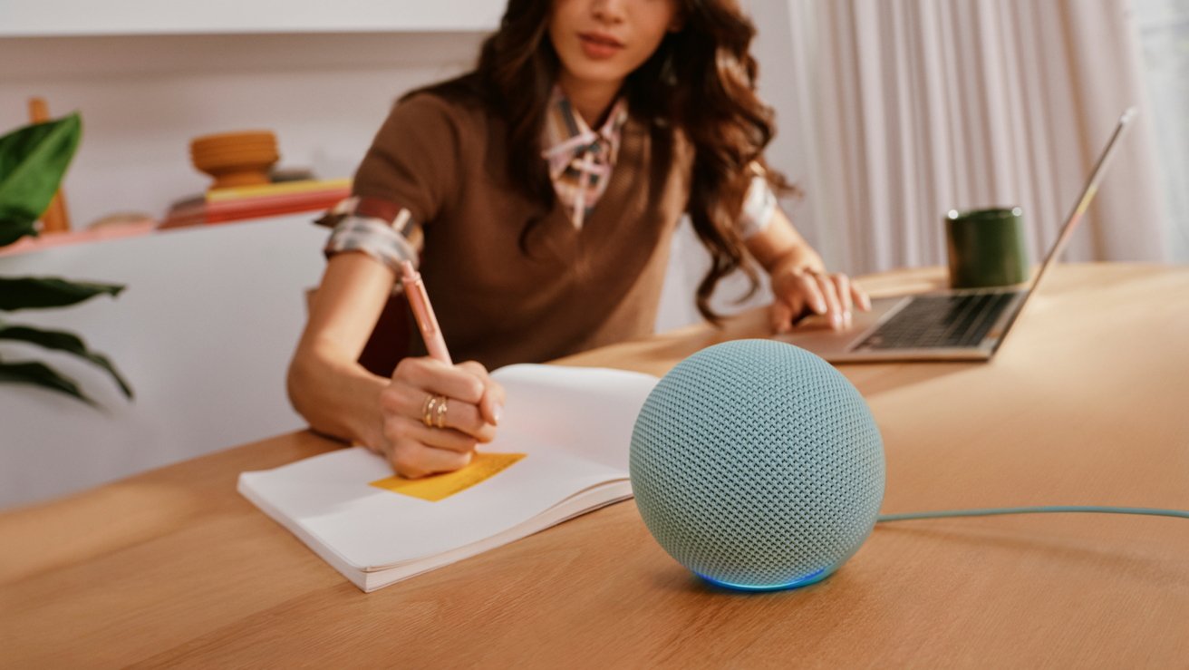 Woman working at a desk with a laptop, writing in a notebook, while a small blue spherical smart speaker sits prominently in the foreground. — Picture from appleinsider.com